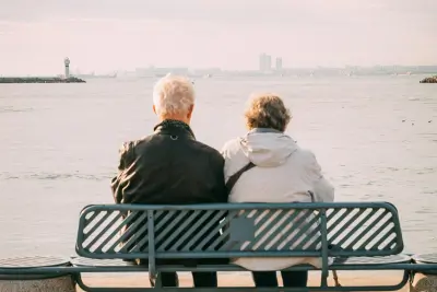 Two individuals seated on a bench overlooking a calm sea, contemplating the horizon, symbolizing planning to calculate retirement.