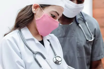 Healthcare professionals in white lab coats with stethoscopes, one with an obscured face and the other wearing a surgical mask.