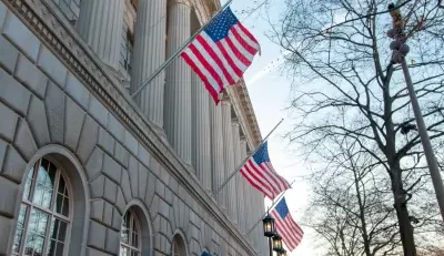 Three American flags waving in front of a classical stone building with large windows and columns, symbolizing stability and tradition, possibly representing an American life insurance company headquarters.