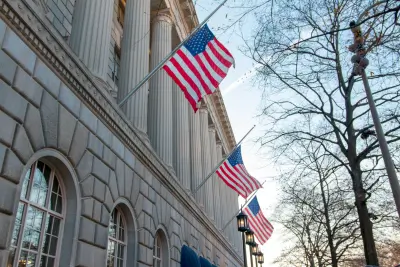 Three American flags waving in front of a classical stone building with large windows and columns, symbolizing stability and tradition, possibly representing an American life insurance company headquarters.