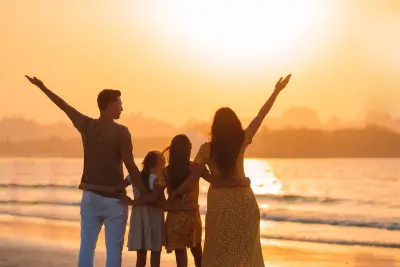 A family of four, with two adults and two children, stands on a beach at sunset, raising their arms in a gesture of joy and freedom, symbolizing the benefits of work-life balance.